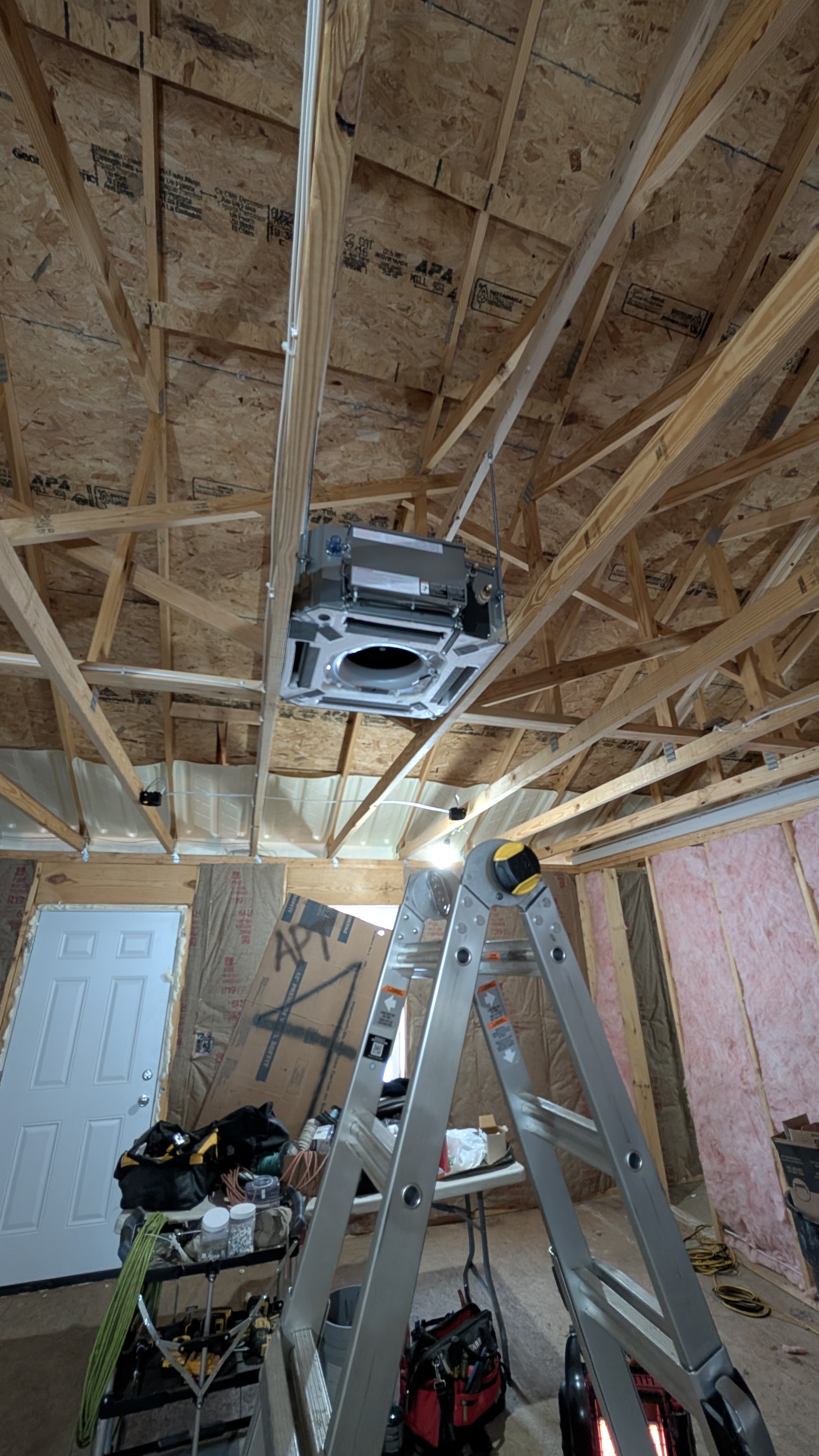 Wide establishing shot of ceiling cassette mini-split install in new-construction outbuilding with tools and ladder