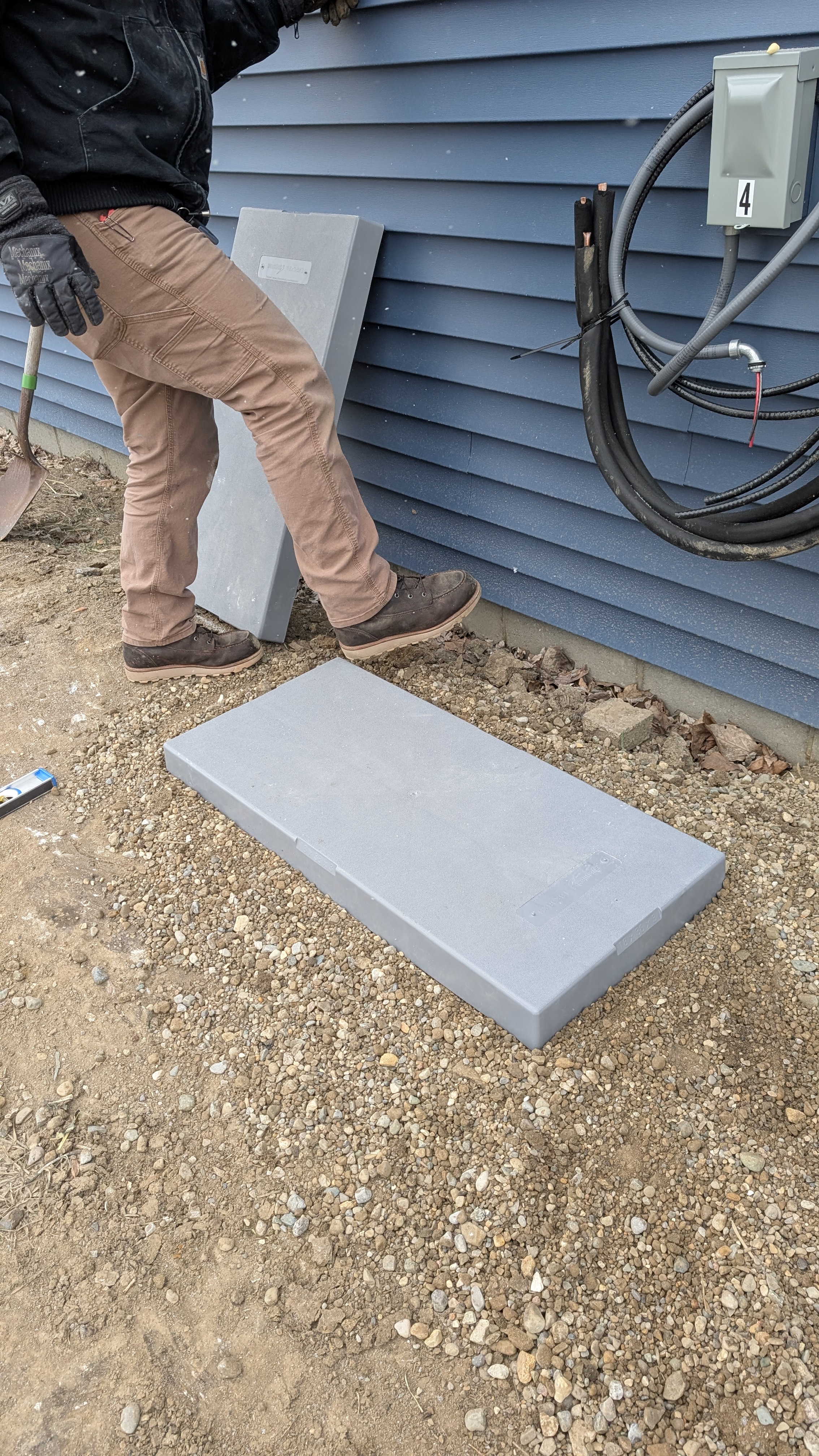 Technician placing concrete condenser pad at new-construction building exterior with disconnect box visible