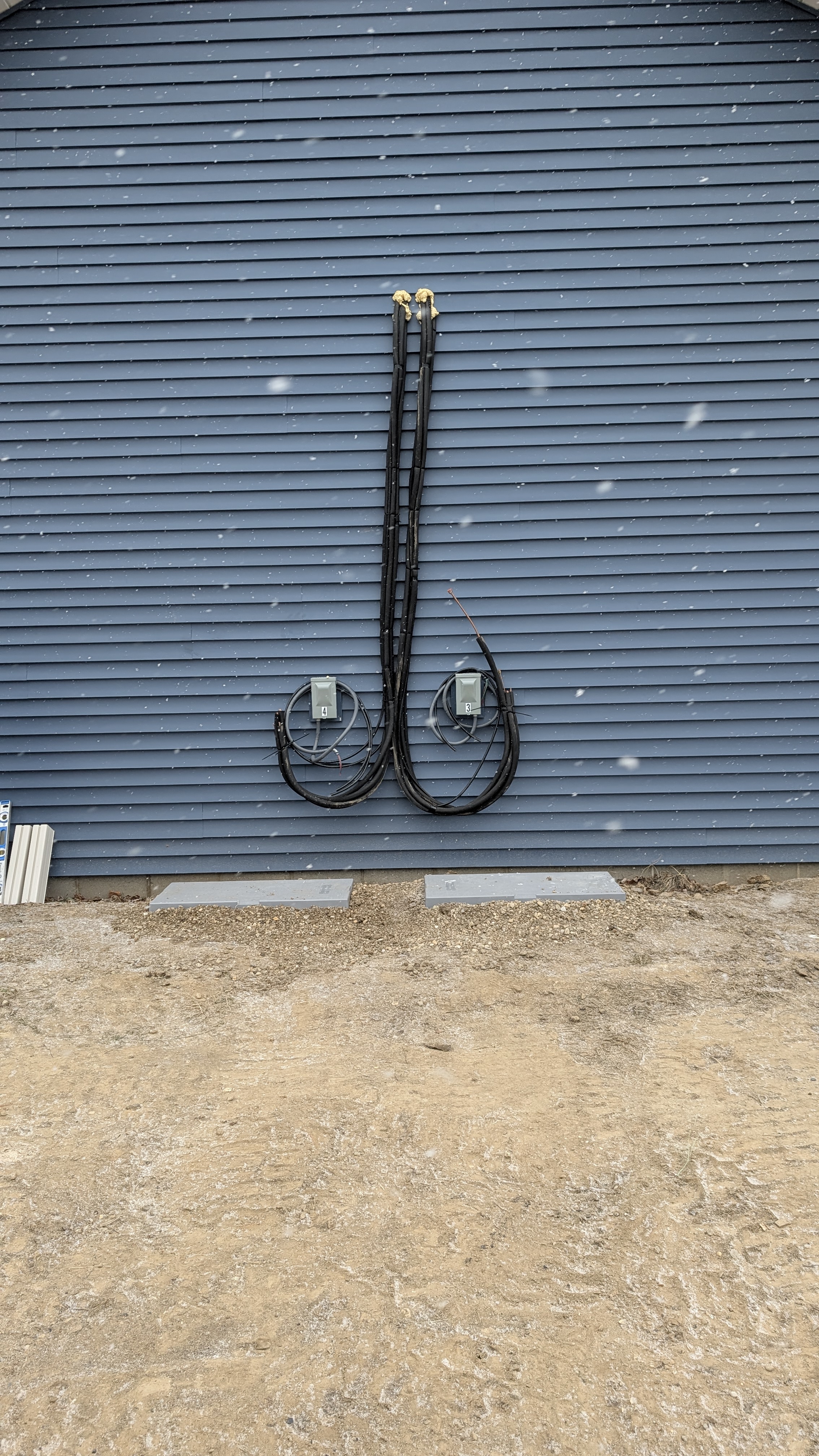 Dual-zone mini-split line-sets and disconnect boxes on exterior wall with condenser pads staged in snow