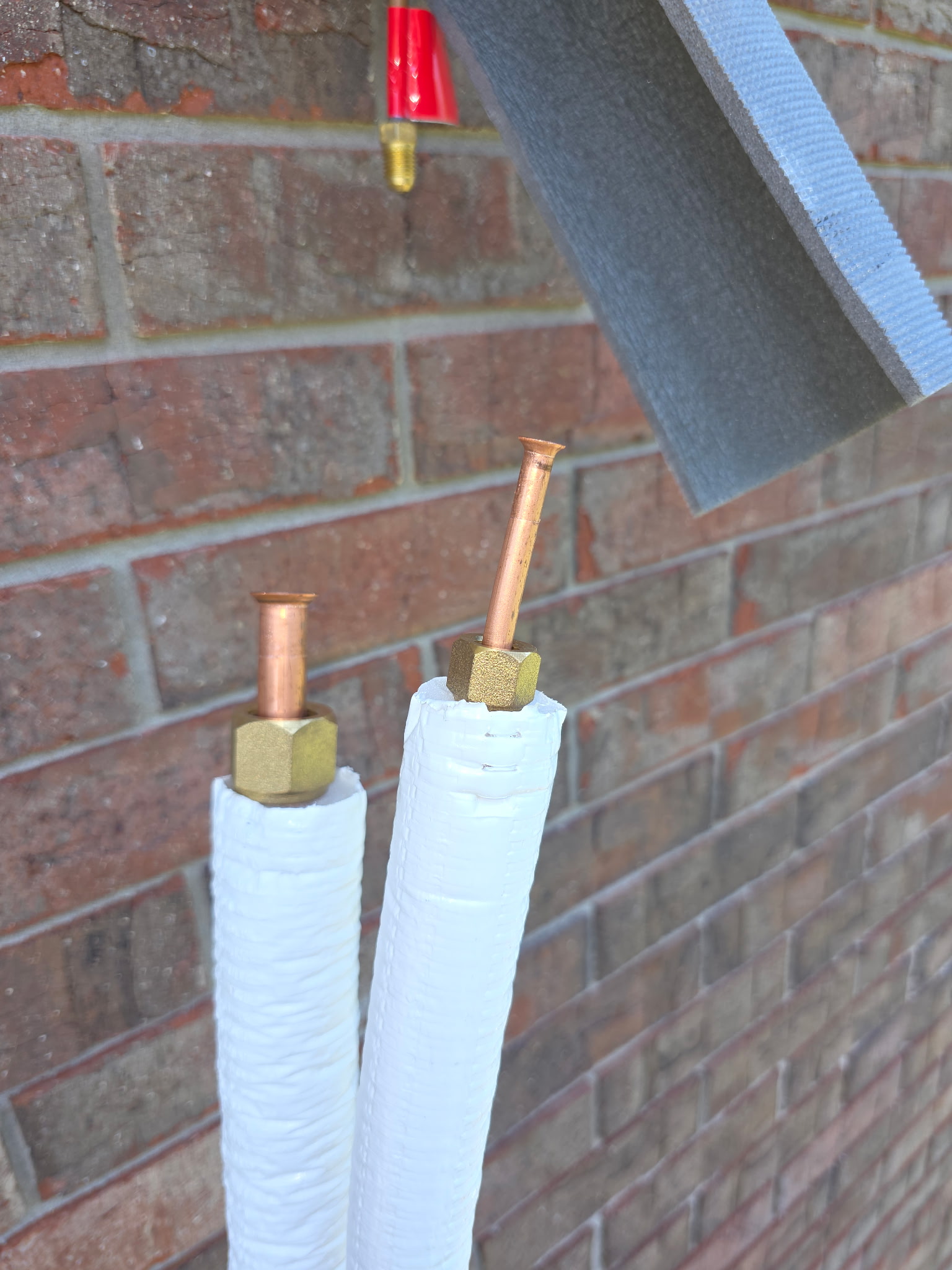 Pair of insulated mini-split refrigerant copper lines with brass flare nuts at brick exterior