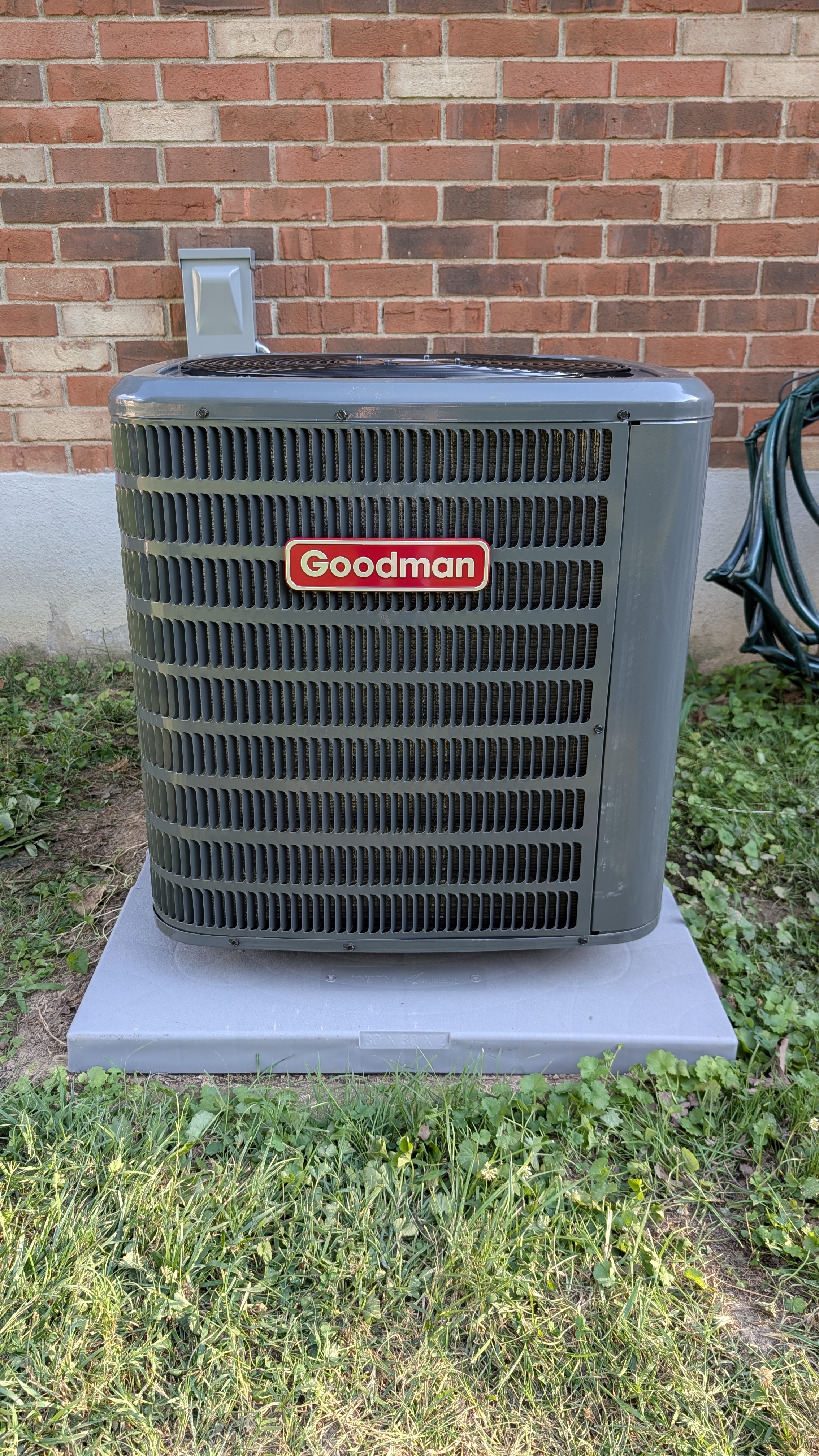 Goodman central AC condenser unit installed on pad outside brick home